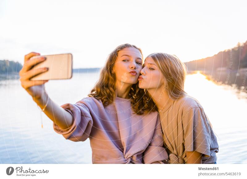 Friends puckering lips while taking selfie on smart phone against lake color image colour image outdoors location shots outdoor shot outdoor shots sunset
