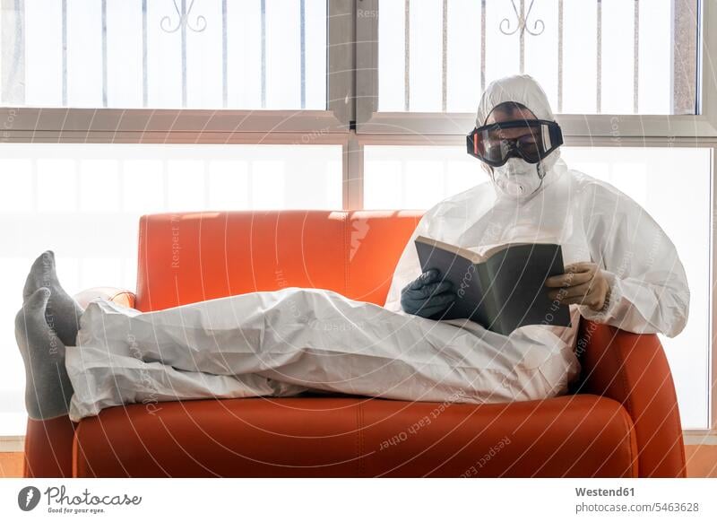 Woman in protective clothes, sitting on couch, reading book books masks couches settee settees sofa sofas Seated White Colors stand at home Ideas protecting
