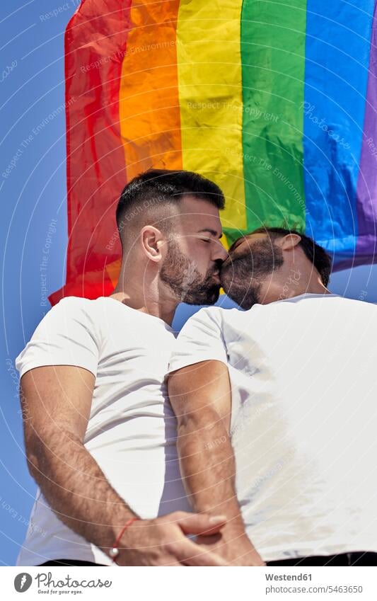 Gay couple kissing while standing under rainbow flag during sunny day color image colour image outdoors location shots outdoor shot outdoor shots low angle view