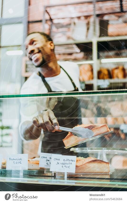 Smiling man working in a bakery taking a piece of cake with a tongs human human being human beings humans person persons African black black ethnicity coloured
