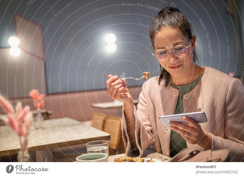 Smiling businesswoman having food while using mobile phone sitting at modern cafe color image colour image indoors indoor shot indoor shots interior