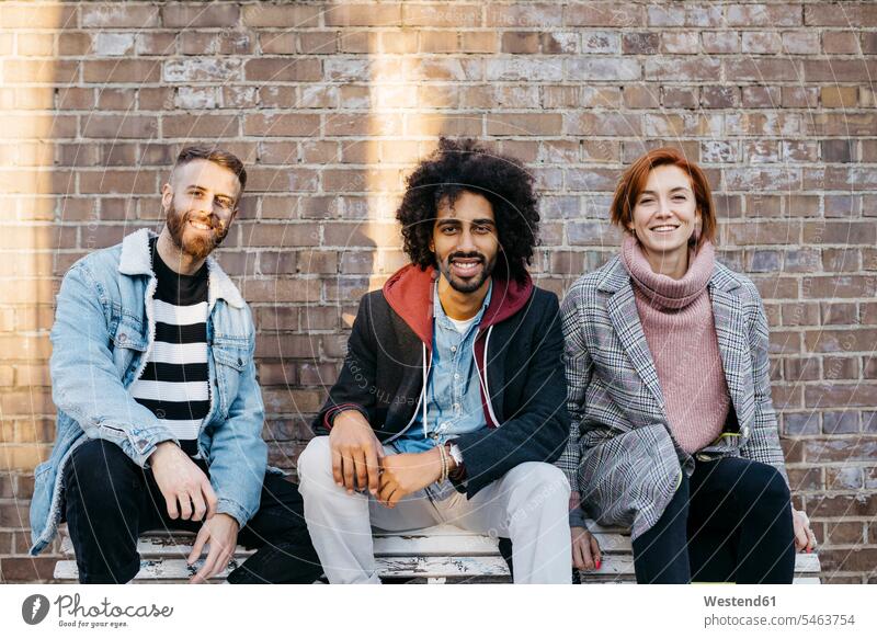 Portrait of three happy friends sitting on a bench in front of a brick wall brick walls happiness portrait portraits Seated benches friendship relationship