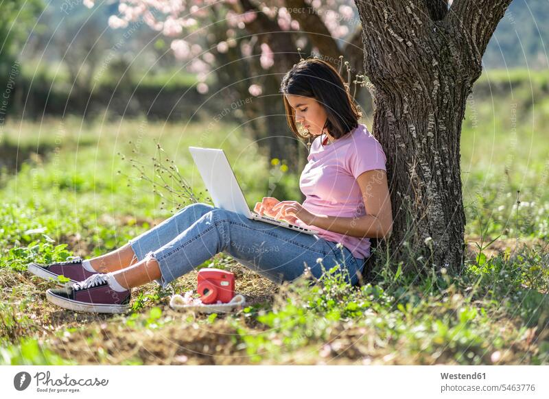 Teenage girl sitting on a meadow leaning against tree trunk using laptop T- Shirt t-shirts tee-shirt computers Laptop Computer Laptop Computers laptops notebook