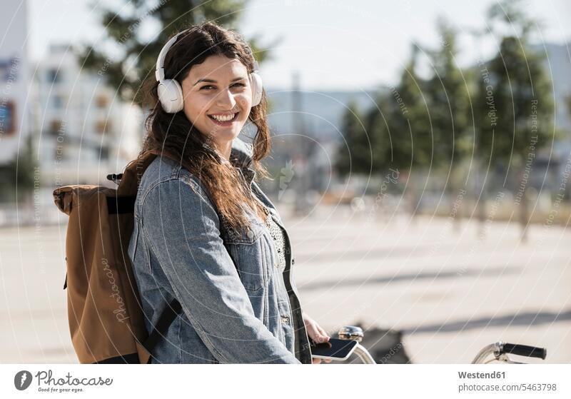 Smiling female student listening music through headphones standing in city on sunny day color image colour image Germany leisure activity leisure activities