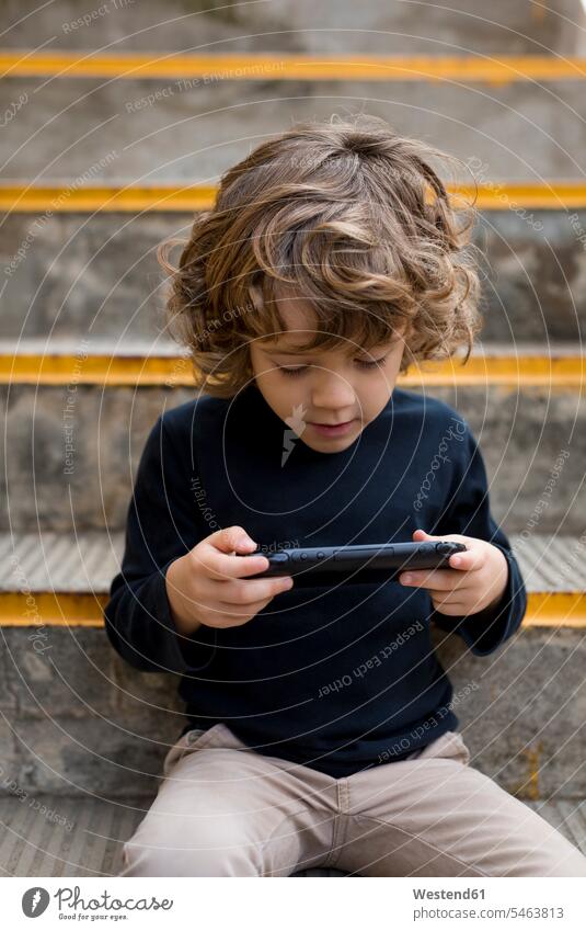 Boy sitting on stairs playing with handheld game console handhelds boy boys males Console stairway Seated child children kid kids people persons human being