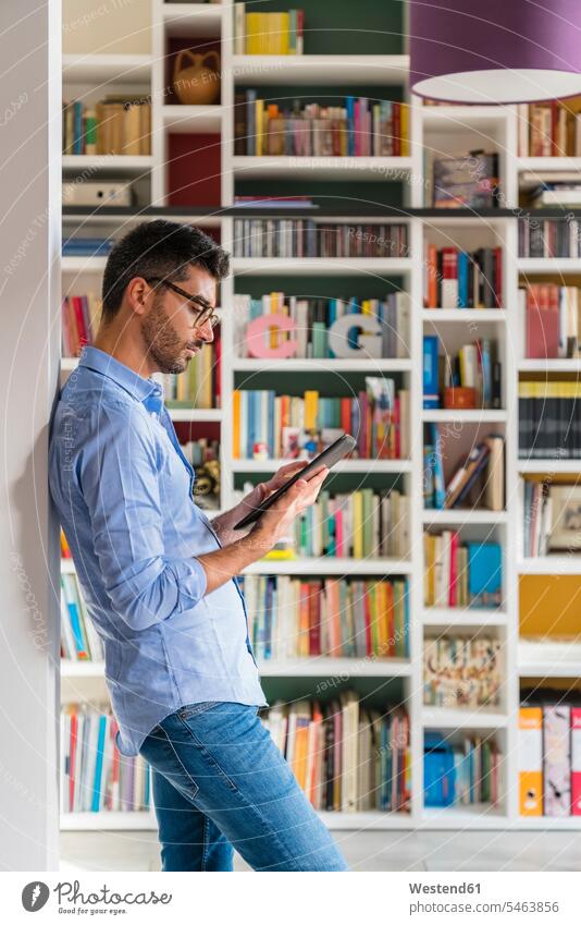 Young man standing in front of bookshelves at home using digital tablet human human being human beings humans person persons caucasian appearance