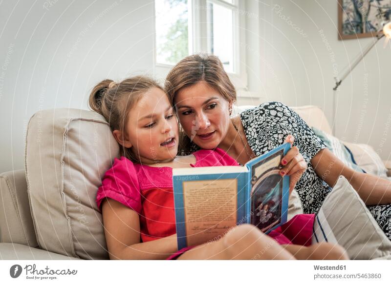 Mother reading book with daughter on couch in living room windows books couches settee settees sofa sofas Seated sit speak speaking talk relax relaxing