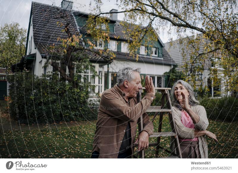 Senior couple with a ladder high fiving in garden of their home relax relaxing smile seasons fall relaxation delight enjoyment Pleasant pleasure Cheerfulness