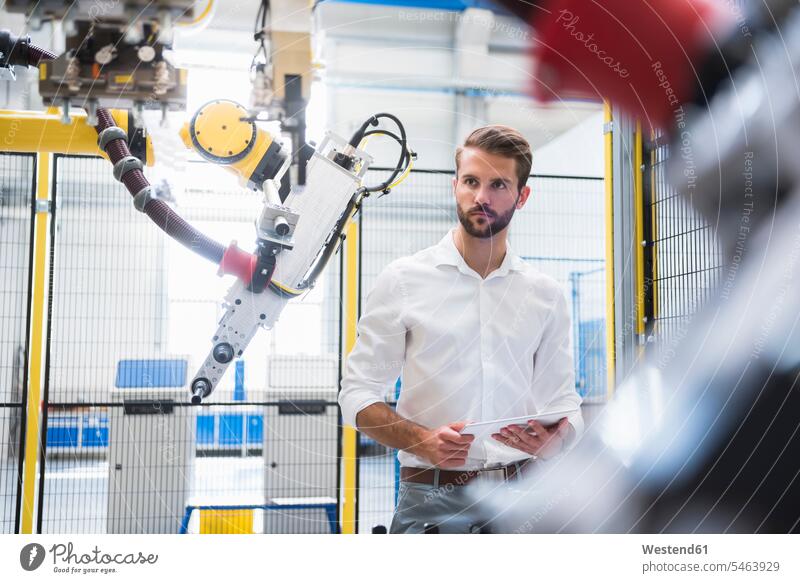 Confident engineer looking at robotic arm while standing with digital tablet in factory color image colour image Germany indoors indoor shot indoor shots