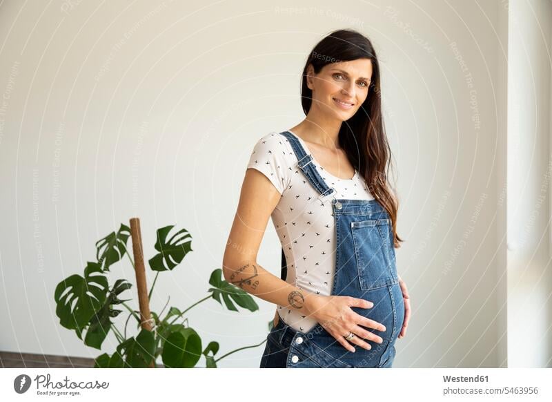 Smiling pregnant woman with hands on stomach standing against wall in new home color image colour image Germany indoors indoor shot indoor shots interior