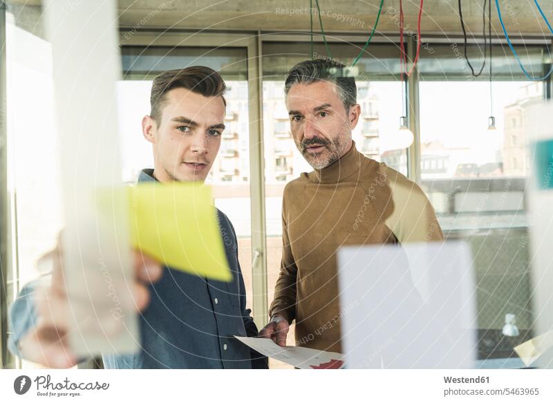 Mature businessman and young man working on adhesive notes at glass pane in office colleague associate associates partner partners partnerships Occupation Work