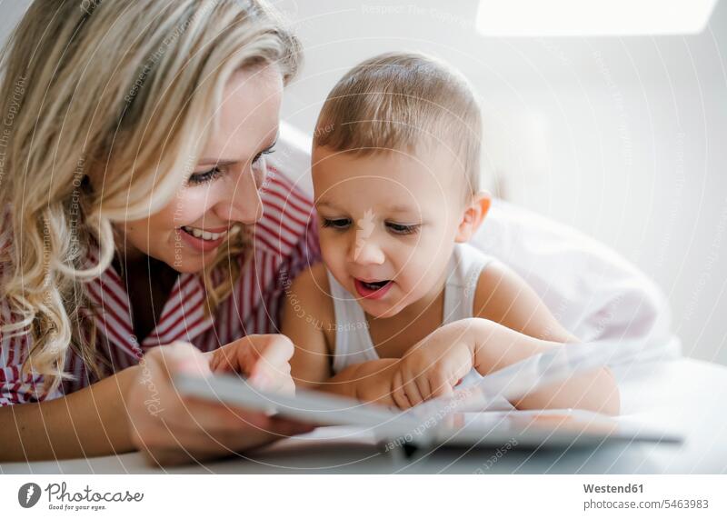 Smiling mother and toddler son lying in bed at home reading a book laying down lie lying down mommy mothers ma mummy mama beds sons manchild manchildren smiling