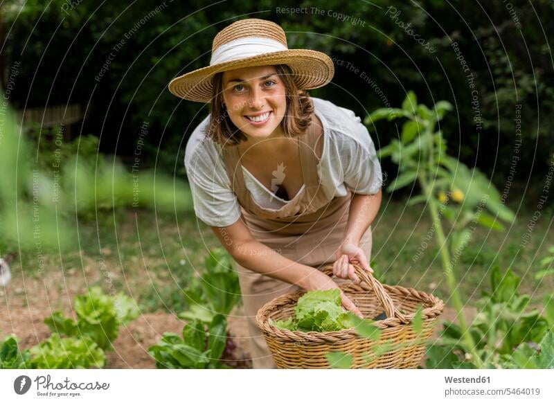 Smiling young woman wearing hat collecting vegetables in wicker basket at yard color image colour image Spain dungarees Bib Overalls Bibs Overall Bibs Overalls