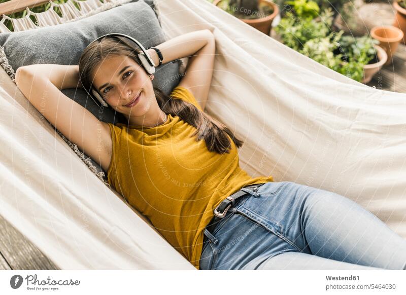 Smiling young woman listening music through headphones while lying on hammock in yard color image colour image Germany leisure activity leisure activities
