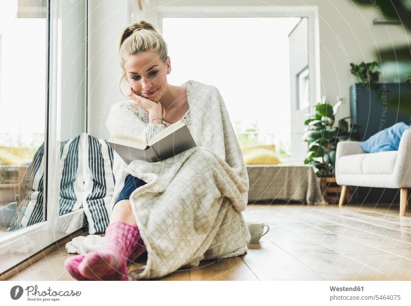 Woman wrapped in a blanket sitting at the window at home reading a book human human being human beings humans person persons celibate celibates singles