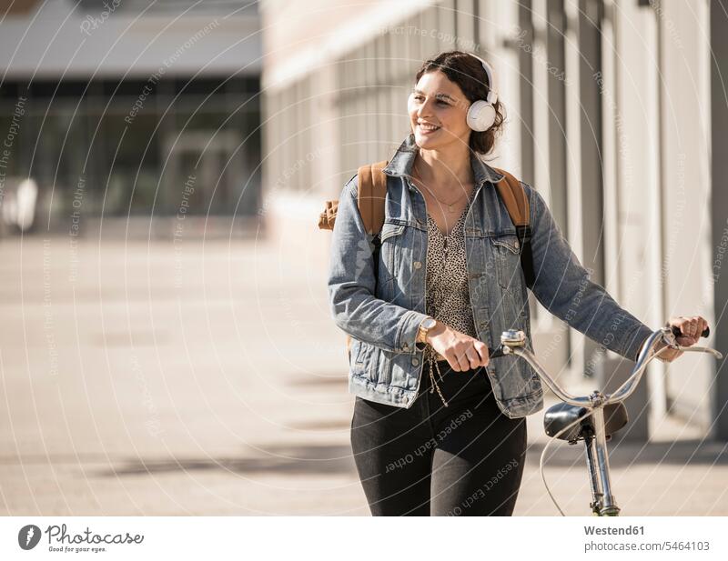 Smiling female commuter listening music while walking with bicycle on street in city color image colour image Germany leisure activity leisure activities