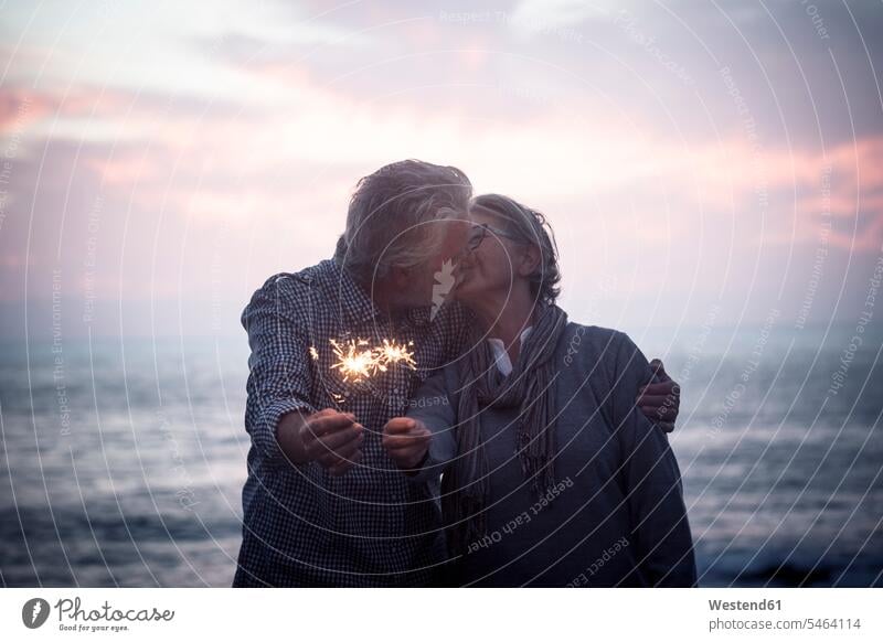 Kissing senior couple standing in front of the sea by sunset holding sparklers Spain evening mood Vignette Moody Sky Romantic Sky sparks togetherness bonding