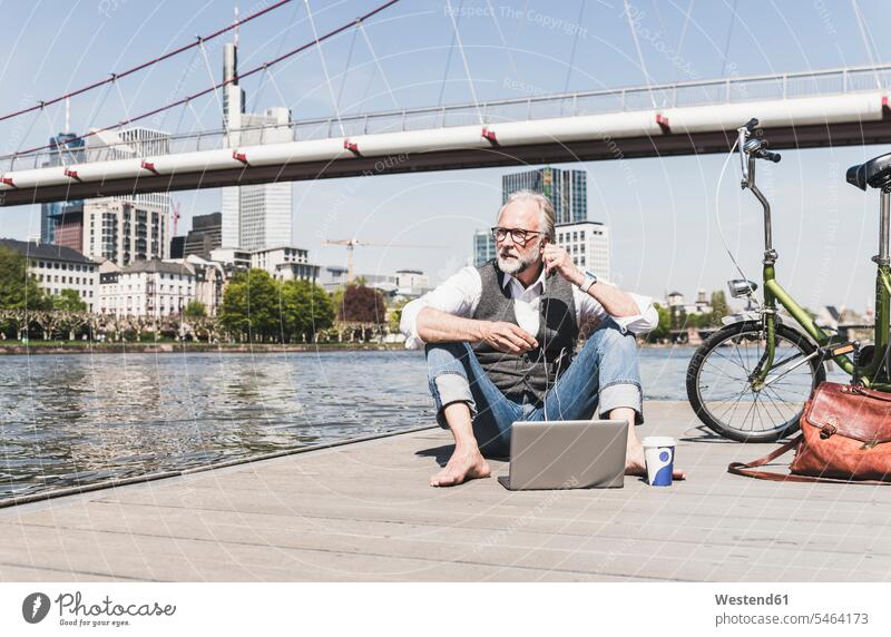 Mature man with laptop, earbuds and bicycle sitting at the riverside in the city town cities towns riverbank bikes bicycles Seated men males Laptop Computers