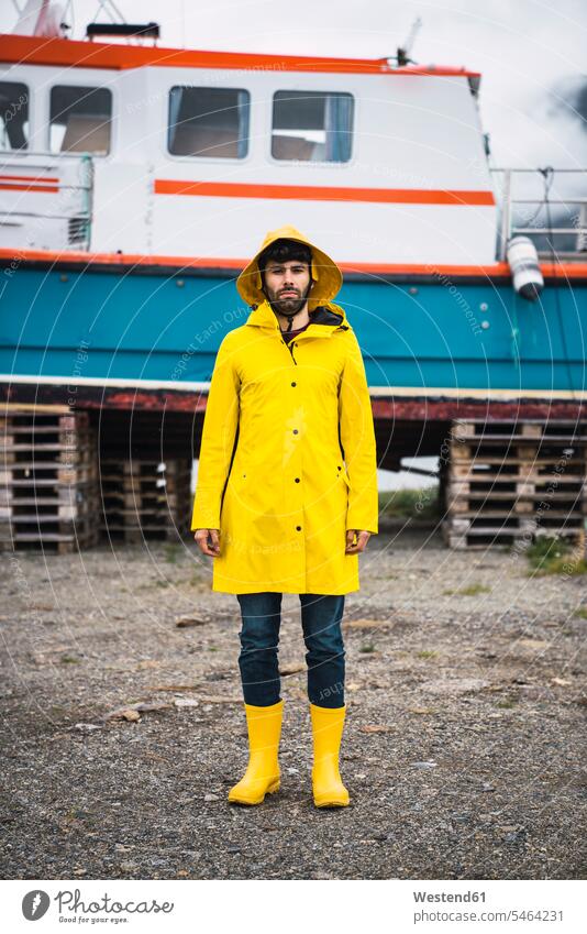 Young man standing in front of a ship, wearing rain clothes, Lapland, Norway rainjacket cagoule rain jacket young man young men Wellington Boot rubber boots