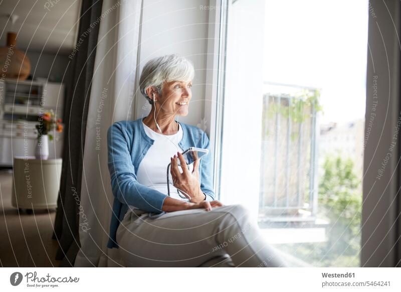 Active senior woman listening to music on smart phone while looking out of window sitting at home color image colour image indoors indoor shot indoor shots