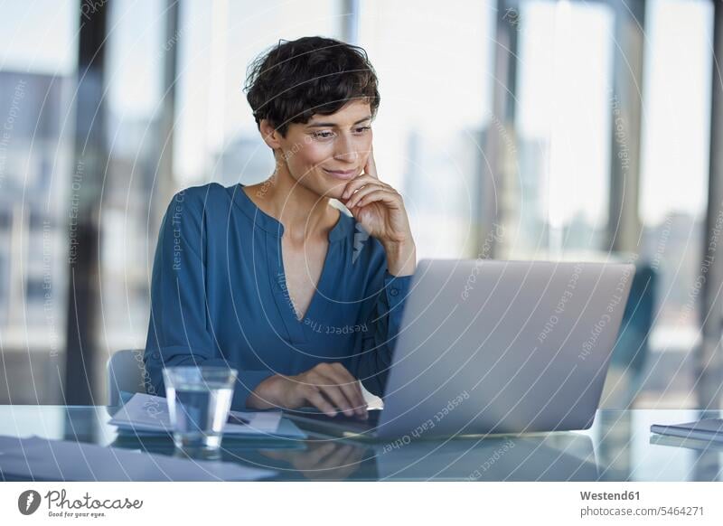 Businesswoman sitting at desk in office using laptop working At Work businesswoman businesswomen business woman business women offices office room office rooms