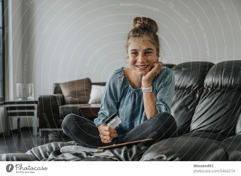 Smiling girl with digital tablet and credit card sitting on sofa at home color image colour image Germany indoors indoor shot indoor shots interior