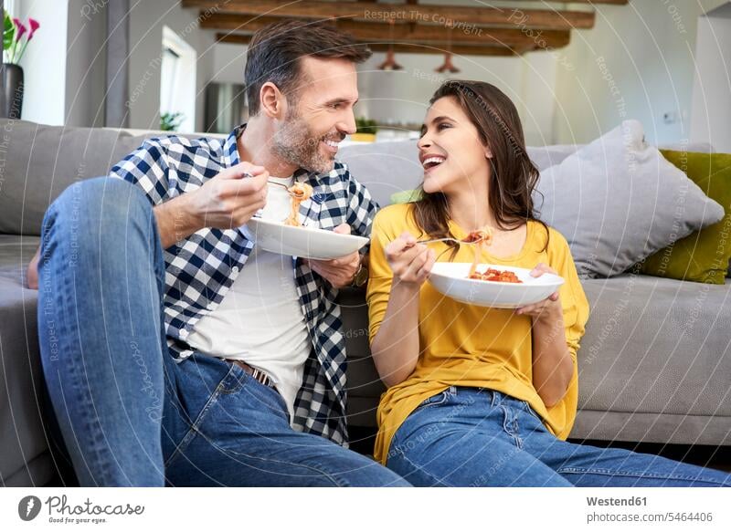 Couple sitting in living room, eating spaghetti Plate dish dishes Plates living rooms livingroom laughing Laughter Spaghetti Seated couple twosomes partnership