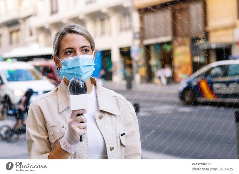 Female journalist wearing mask with microphone looking away while standing on street in city color image colour image Spain outdoors location shots outdoor shot