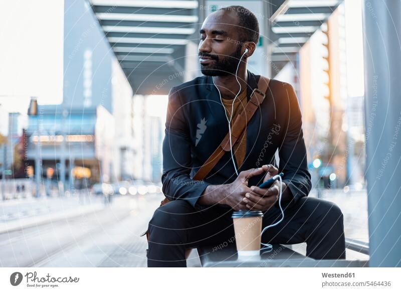Young businessman with coffee to go listening music with earphones and smartphone relaxing at tram stop human human being human beings humans person persons