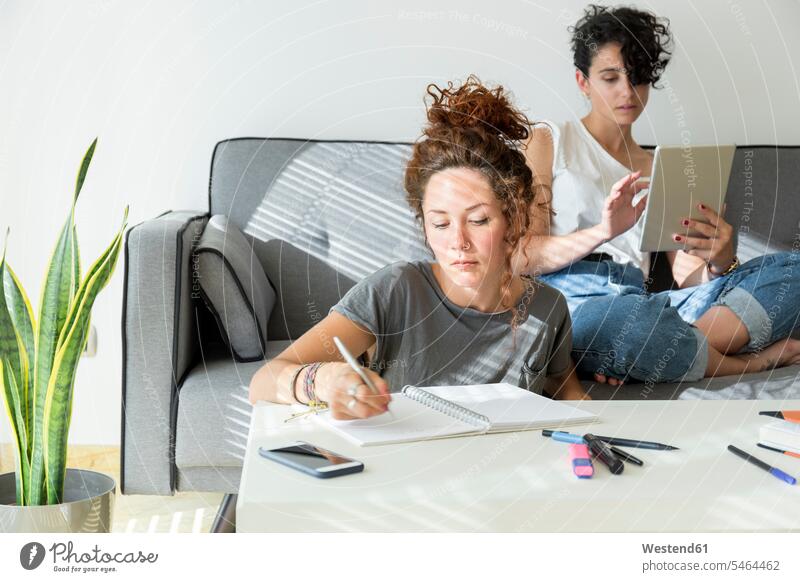 Young woman taking notes at home with friend sitting on couch using tablet human human being human beings humans person persons caucasian appearance