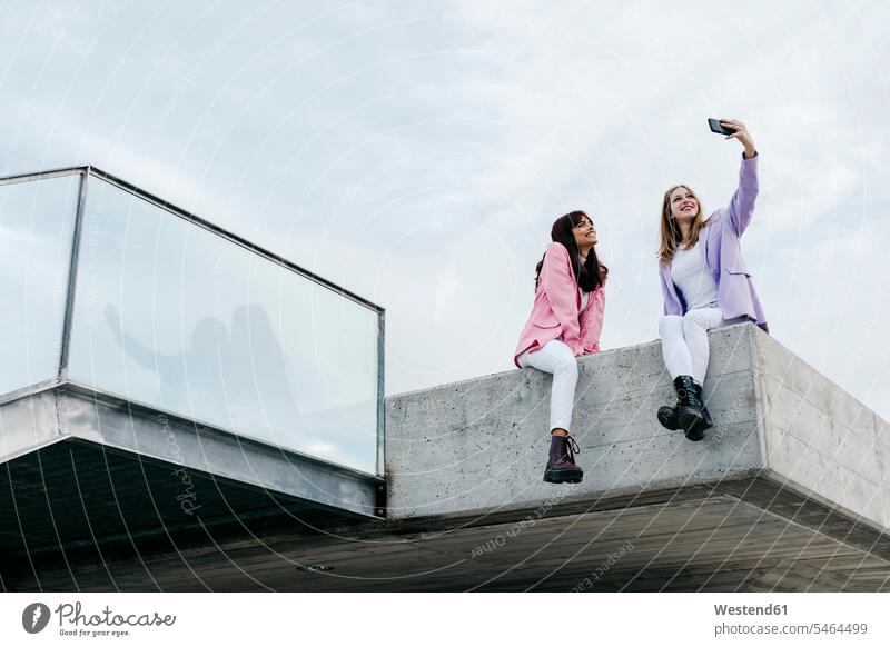 Sisters taking selfie through smart phone while sitting on rooftop in city against sky color image colour image outdoors location shots outdoor shot
