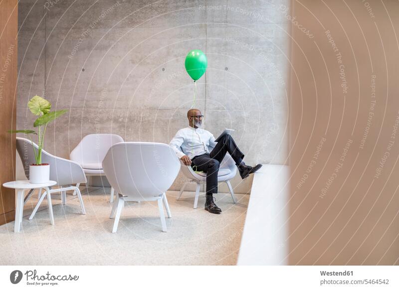 Mature businessman with green balloon sitting on armchair using digital tablet Businessman Business man Businessmen Business men Seated use balloons Arm Chairs