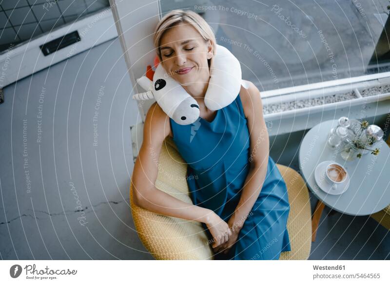 Smiling woman sitting in chair, taking a nap with unicorn pillow around her neck human human being human beings humans person persons caucasian appearance