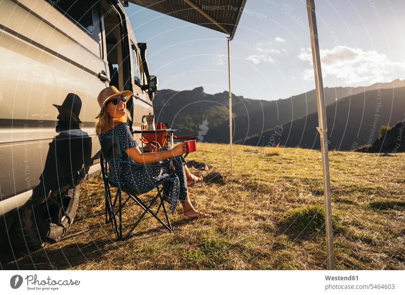 Happy woman in sunglasses with coffee cup laughing while sitting by motor home during vacations color image colour image outdoors location shots outdoor shot