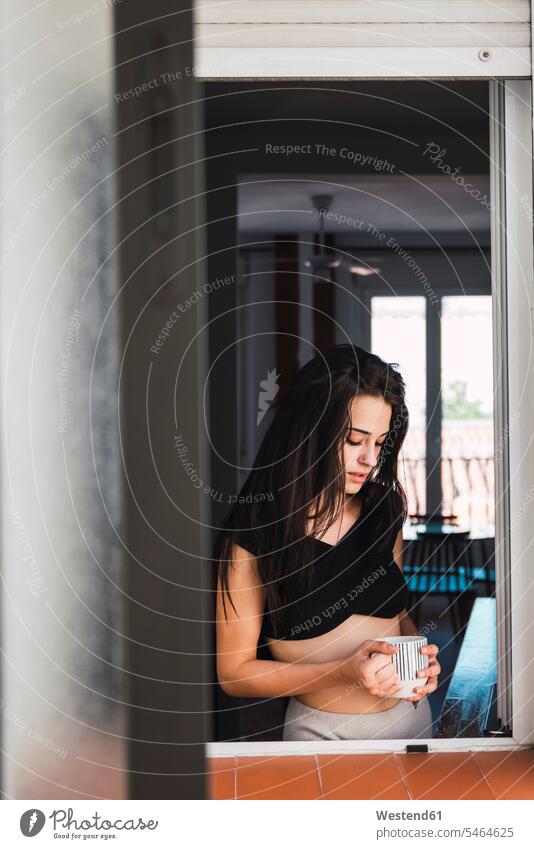 Serious beautiful young woman with cup of coffee at the window at home Coffee females women Coffee Cup Coffee Cups serious earnest Seriousness austere windows