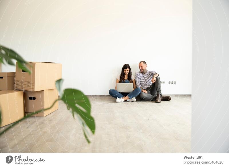 Man sitting with wife using laptop on hardwood floor against wall in new unfurnished home color image colour image Germany indoors indoor shot indoor shots