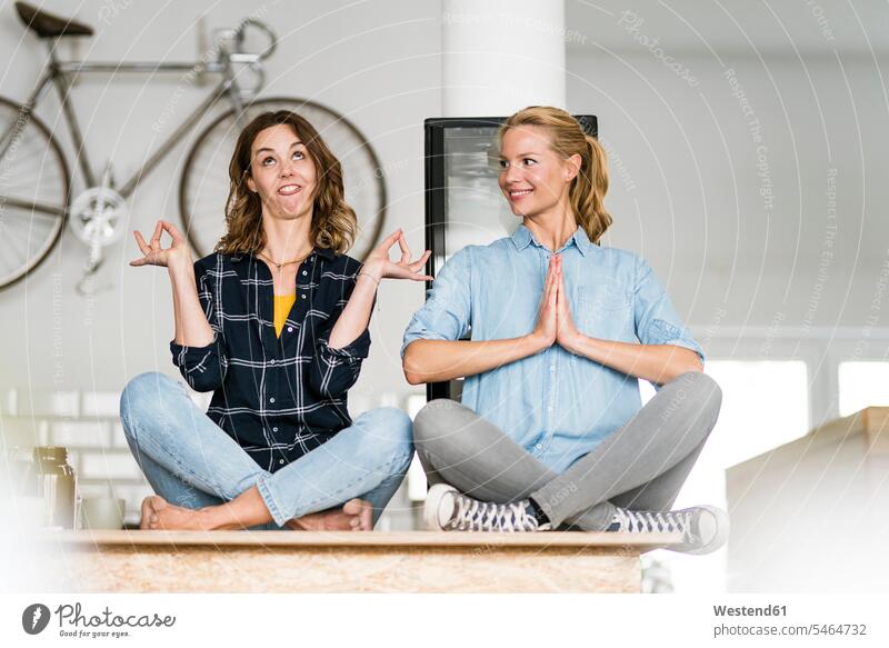 Two young women sitting cross-legged on the counter of their coffee shop, one meditating , other pulling funny faces human human being human beings humans