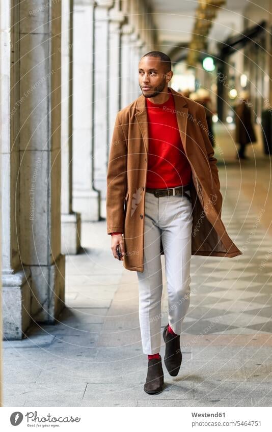 Fashionable young man wearing red pullover and brown coat walking along arcade sweater jumper Sweaters arcades fashionable men males Coat Coats going Adults