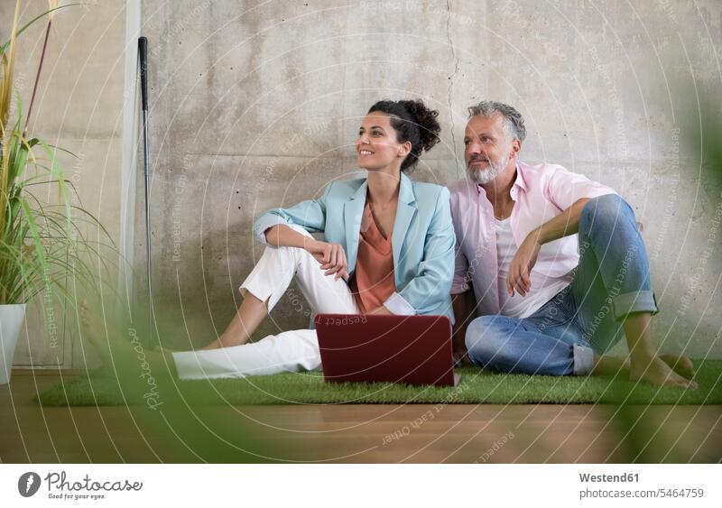 Casual businessman and businesswoman sitting on artificial turf in a loft sharing laptop lofts Businessman Business man Businessmen Business men businesswomen