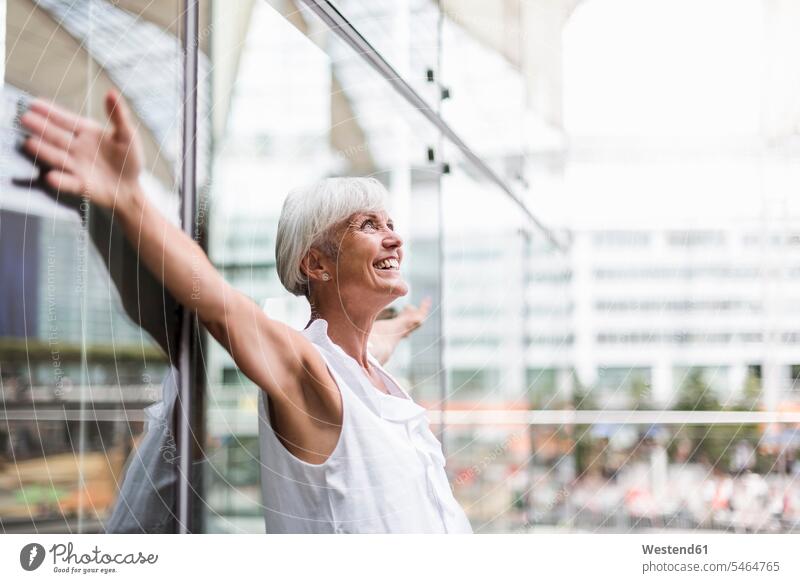 Happy senior woman leaning against glass facade with outstretched arms senior women elder women elder woman old portrait portraits happiness happy Glass front