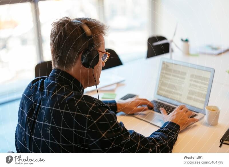 Businessman wearing headphones using laptop at desk in office Laptop Computers laptops notebook Business man Businessmen Business men desks headset offices