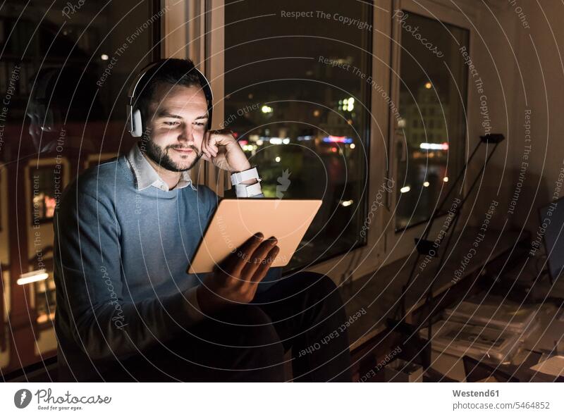 Businessman sitting on window sill in office at night using tablet and headphones Business man Businessmen Business men headset use by night nite