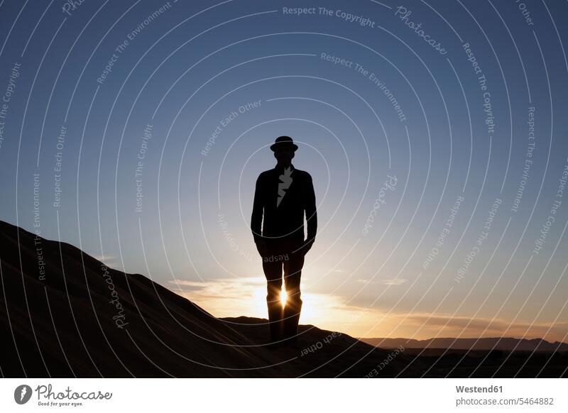 Morocco, Merzouga, Erg Chebbi, silhouette of man wearing a bowler hat standing on desert dune at sunset sunsets sundown men males desert dunes silhouettes hats