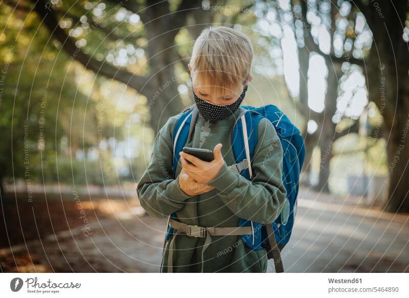 Boy text messaging on smart phone wearing protective face mask while standing in public park color image colour image outdoors location shots outdoor shot
