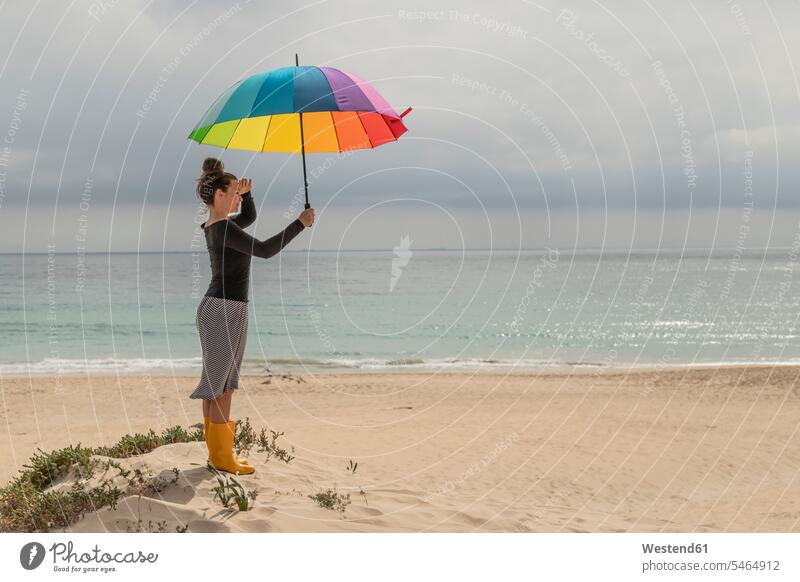 Woman with colorful umbrella sitting on the beach prismatic colours Rainbow Color prismatic colors Tolerance tolerant tolerantly Wellington Boot rubber boots