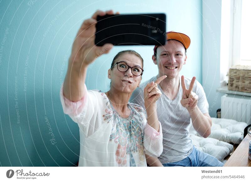 Mother and son sitting at the kitchen table, taking smarrtphone selfies Germany Visit Selfie Selfies baseball cap visor cap peaked cap visored cap visiting