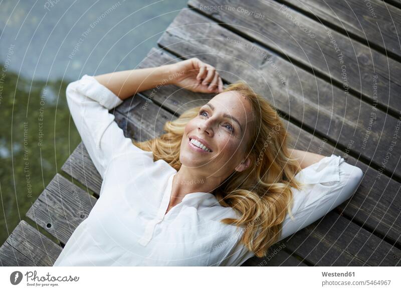 Smiling blond woman lying on wooden jetty at a lake blond hair blonde hair jetties laying down lie lying down females women lakes smiling smile people persons
