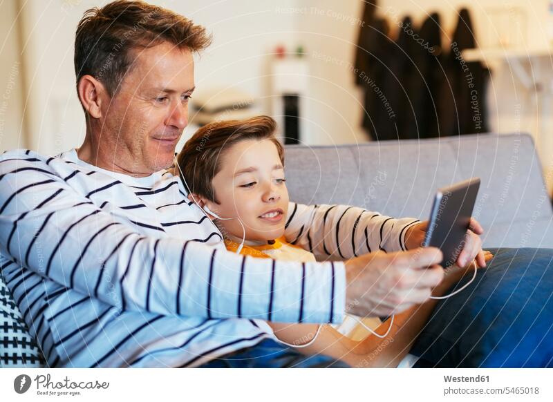 Father and son with earbuds and tablet on couch at home father pa fathers daddy dads papa sons manchild manchildren earphones ear phone ear phones settee sofa