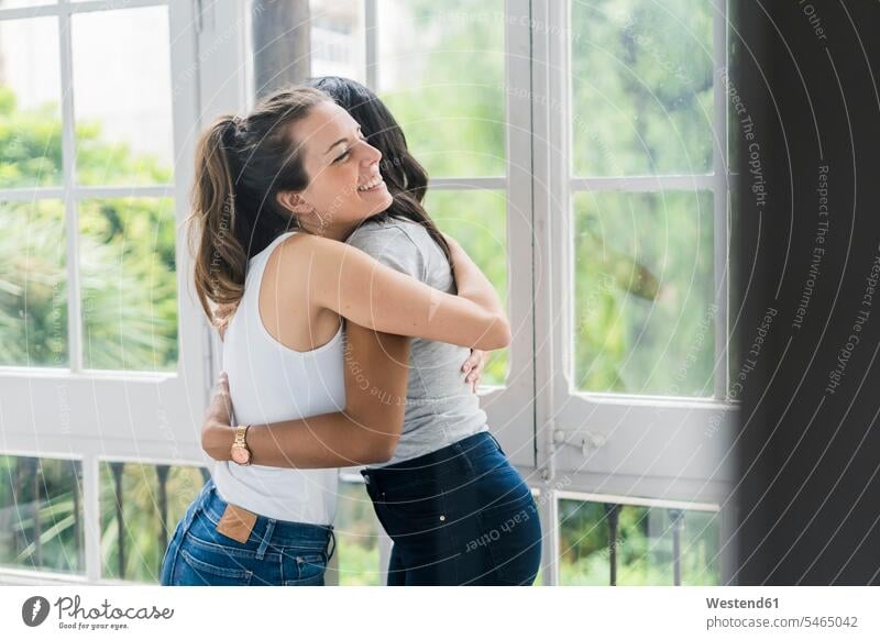 Two happy girlfriends hugging at the window embracing embrace Embracement female friends woman females women happiness windows mate friendship Adults grown-ups