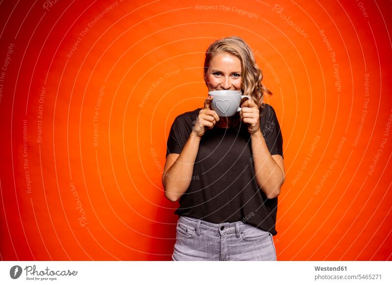 Happy woman holding coffee cup with heart shape while standing against orange background color image colour image coloured background colored background